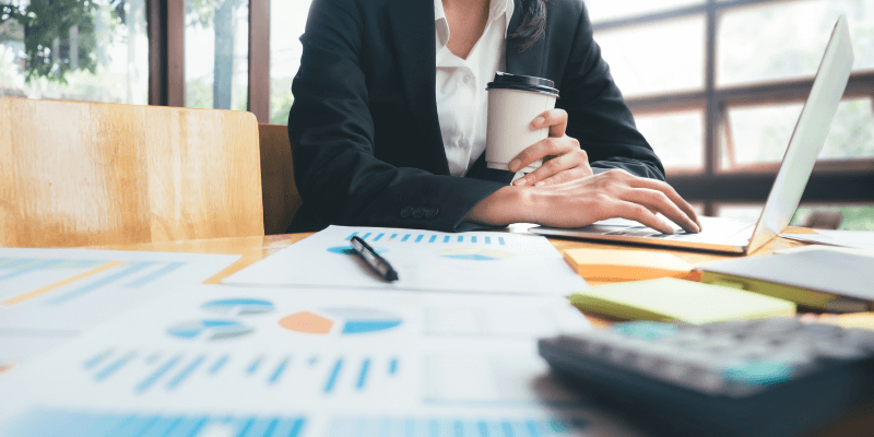 Woman drinking coffee while working on her laptop. Her desk is covered in budgets and spreadsheets.