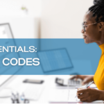 Woman in a yellow shirt and glasses working on her office computer. Text overlay read “EDI Essentials: Top 10 Codes”