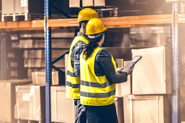 2 workers in yellow reflective gear in a warehouse looking at a tablet.