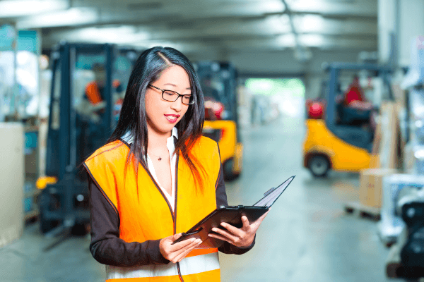 Woman in an orange reflective jacket examining a tablet. She is in a warehouse with forklifts behind her.