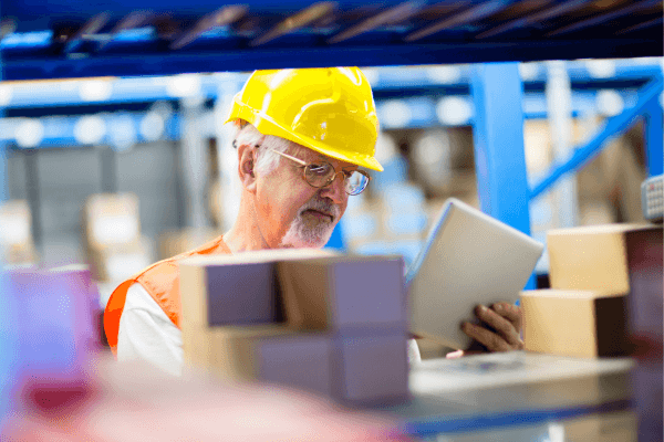 Older man wearing safety gear, sorting boxes in a warehouse while consulting a handheld tablet.