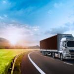 Semi truck driving on a black road across green countryside with blue sky and the sun coming up behind mountains in the distance.