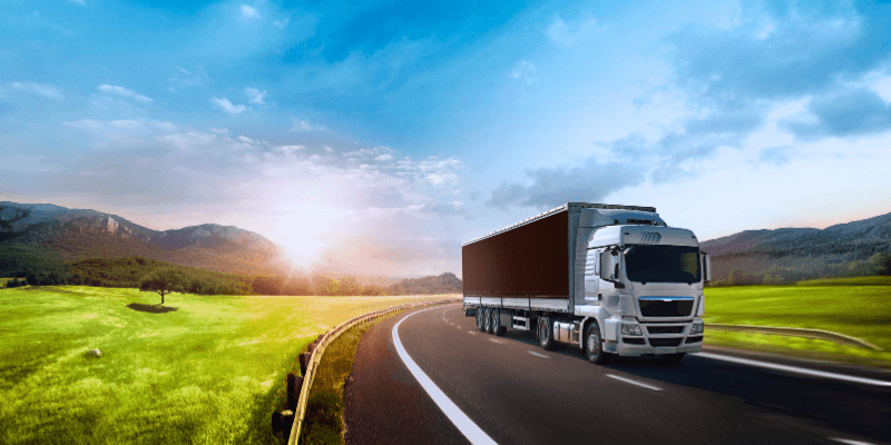 Semi truck driving on a black road across green countryside with blue sky and the sun coming up behind mountains in the distance.